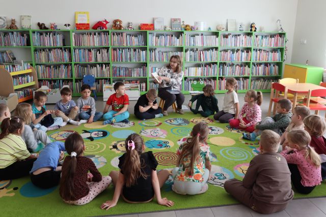 przedszkolaki siedzą na kolorowym dywanie, wśród nich siedzi bibliotekarka z książką. w tle regały z książkami