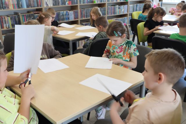 przedszkolaki siedzą przy stolikach, w rękach mają nożyczki, na stołach leżą białe kartki