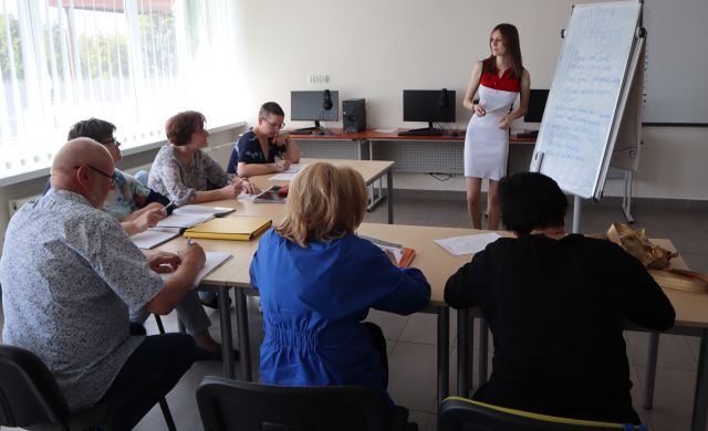 seniorzy siedzą przy stolikach, na stolikach leżą zeszyty i długopisy, przed seniorami stoi kobieta przy flipcharcie