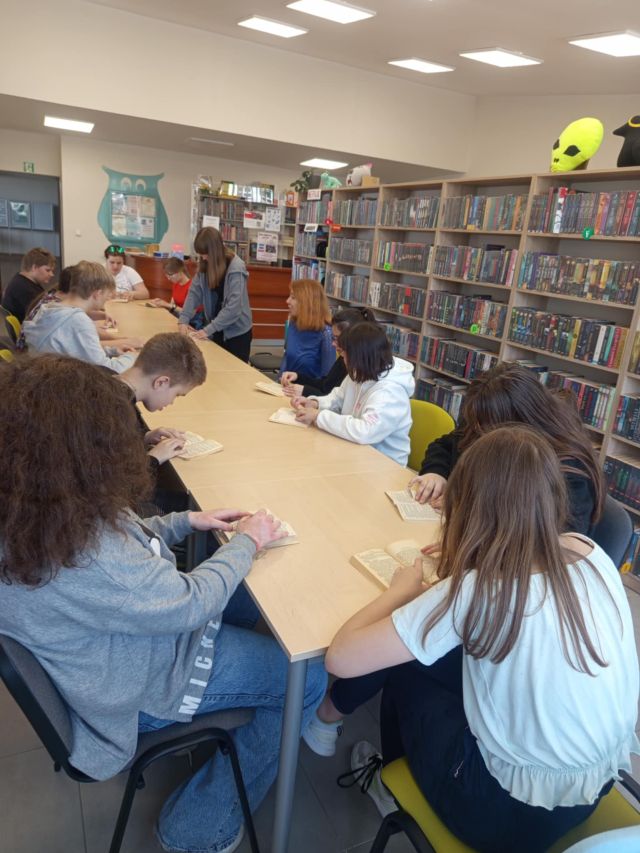 uczniowie siedzą przy stołach, za ich plecami regały z książkami