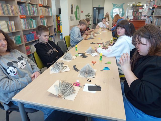uczniowie siedzą przy stołąch i wycinają, składają i malują
