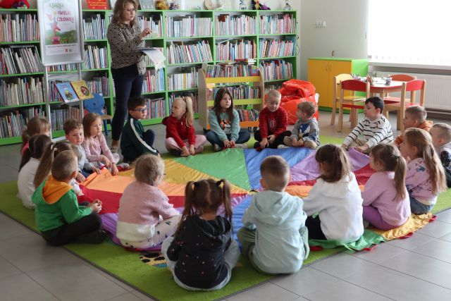 przedszkolaki siedzą na kolorowej  chuście animacyjnej, w tle regały z książkami