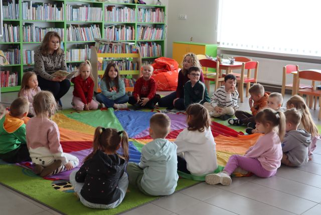 przedszkolaki siedzą na kolorowej  chuście animacyjnej, w tle regały z książkami
