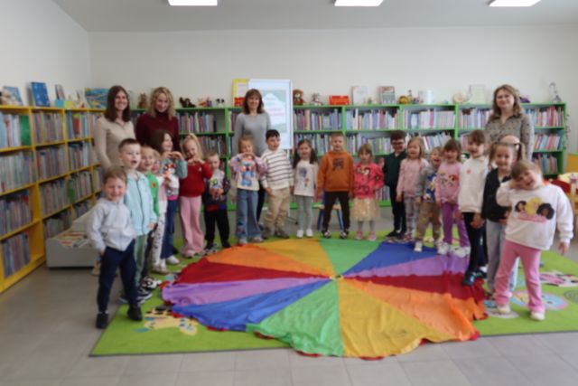 przedszkolaki, dwie naucycielki i bibliotekarki stoją wokół chusty animacyjnej i pozują do zdjęcia., w tle  regały z książkami