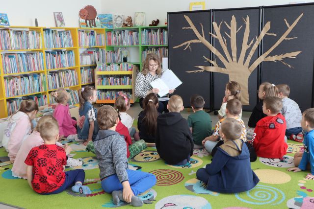 bibliotekarka z książką siedzi na krzesełku przed siedzącą na kolorowym dywanie bibliotekarką, w tle regały z książkami