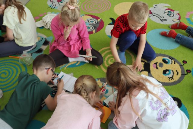 dzieci siedzą na kolorowym dywanie i malują na kartkach