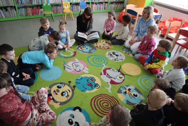przedszkolaki i bibliotekarka z książką siedzą na kolorowym dywanie
