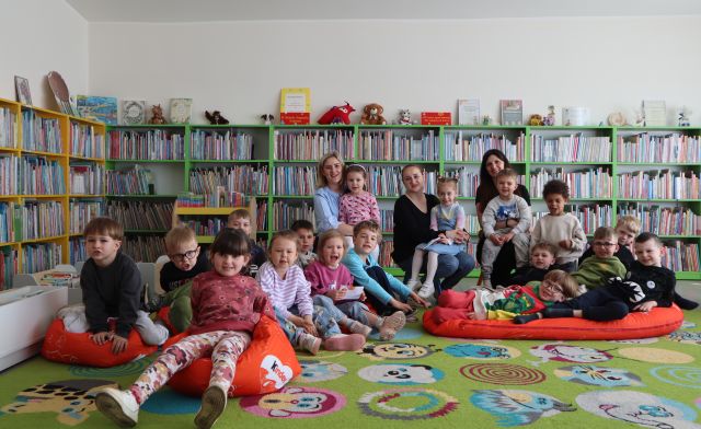 przedszkolaki i nauczycielki siedzą na kolorowym dywanie i pufach i pozują do zdjęcia, w tle regały z książkami