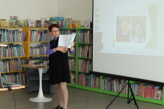 kobieta z książką, w tle regały z książkami i ekran