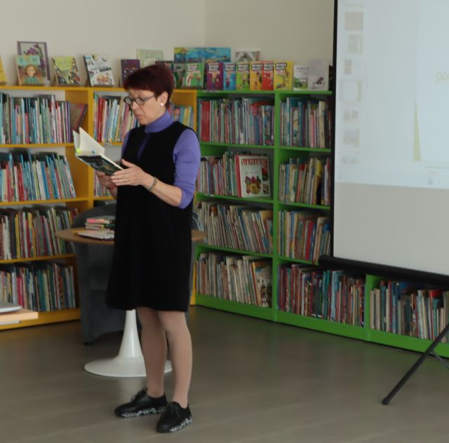 kobieta czyta książkę, w tle regały z książkami i ekran