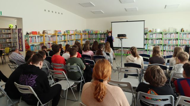 dzieci uczestniczące w spotkaniu z autorką siedzą na krzesłach, w tle autorka, regały z książkami, ekran