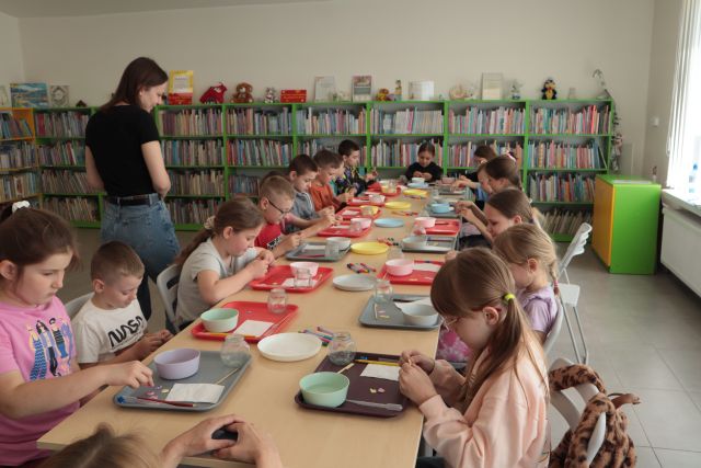 dzieci siedzą przy stołach, na stołach tacki, miski, w tle regały z książkami