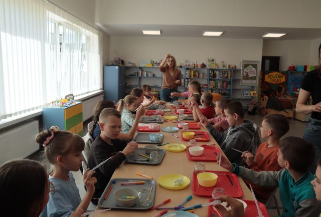 20 dzieci siedzą przy połączonych w rząd stołach i robią robaczki