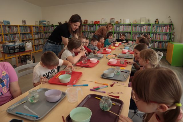 dzieci siedzą przy stołach na warsztatach, obok stoi kobieta wlewa płyn do misek