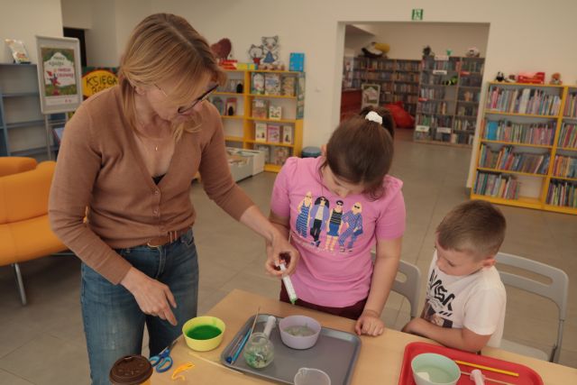 kobieta strzykawką wstrzykuje kolorową ciecz do miseczki, obok stoi dziewczynka i chłopiec