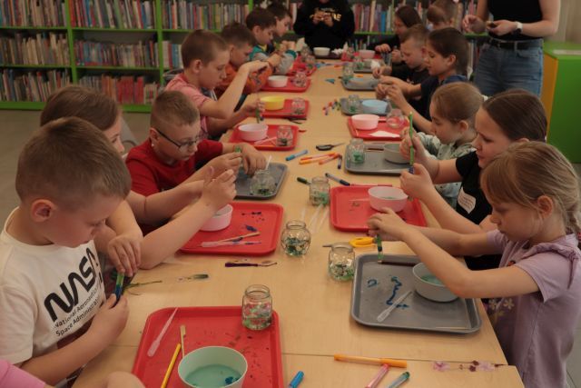 dzieci uczestniczące w warsztatach siedzą przy stołach i pracują, w tle regały z książkami