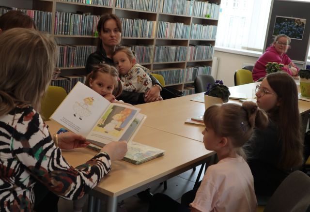 bibliotekarka z książką czyta dzieciom i rodzicom siedzącym przy rzędzie stołów