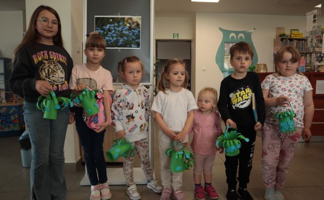 7 dzieci pozuje do zdjęcia z wykonanymi przez siebie zielonymi koszyczkami z papieru i naklejonymi na nie niebieskimi niezapominajkami, przestrzeń biblioteczna