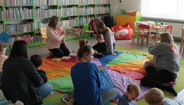 dzieci z mamami siedzą na kolorowej chuście animacyjnej, wśród nich siedzi bibliotekarka z książką
