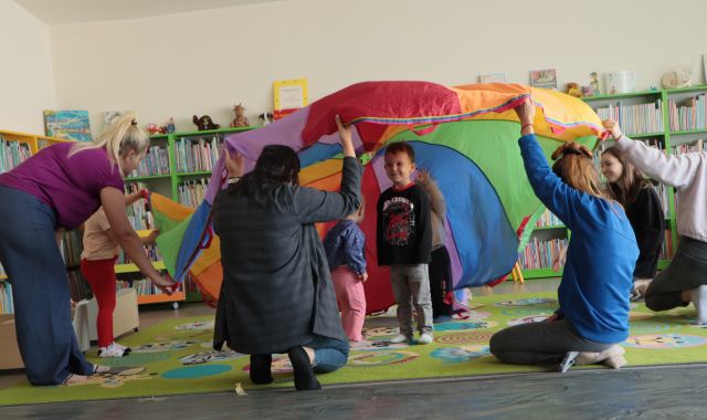 rodzice trzymają chustę animacyjną, pod chustą małę dzieci, w tle regały z książkami