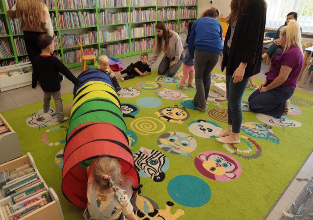 dzieci przechodzą przez tunel animacyjny, pozostałę dzieci z mamami stoją i siedzą na kolorowym dywanie, w tle regały z książkami