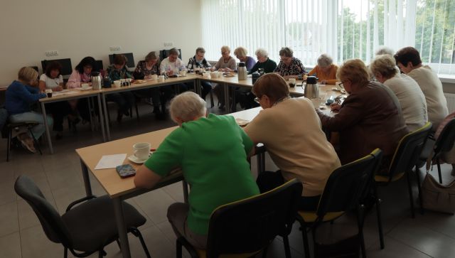 18 seniorek siedzi przy ustawionych w podkowę i pisze na kartkach