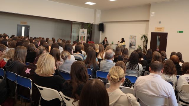 przeszło 200 osób siedzi na krzesłąch, przed nimi w tle siedzą dwie młode kobiety