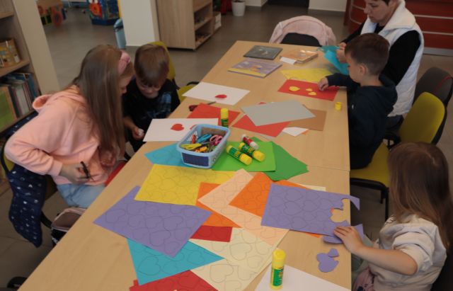 4 dzieci i dwie mamy siedzą przy stołach, na stołąch kolorowe kartki, dzieci mają w rękach nożyczki