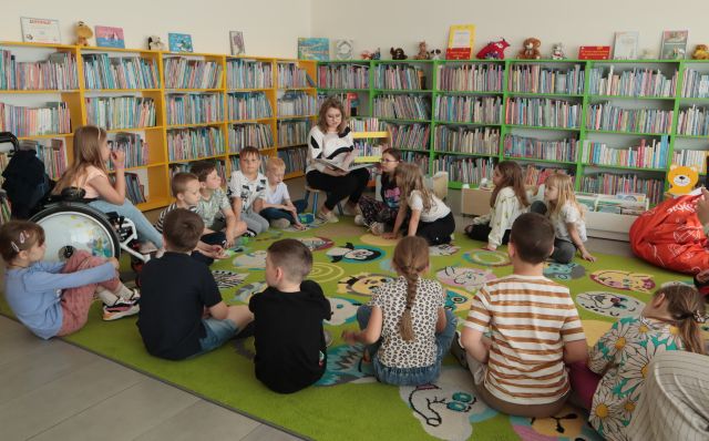 bibliotekarka siedzi z książką na krześle, na dywanie siedzą dzieci, w tle regały z książkami