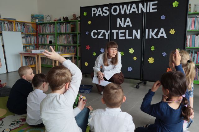 bibliotekarka kuca przed siedzącymi na dywanie dziećmi, w tle tablica czarna z napisem Pasowanie na Czytelnika