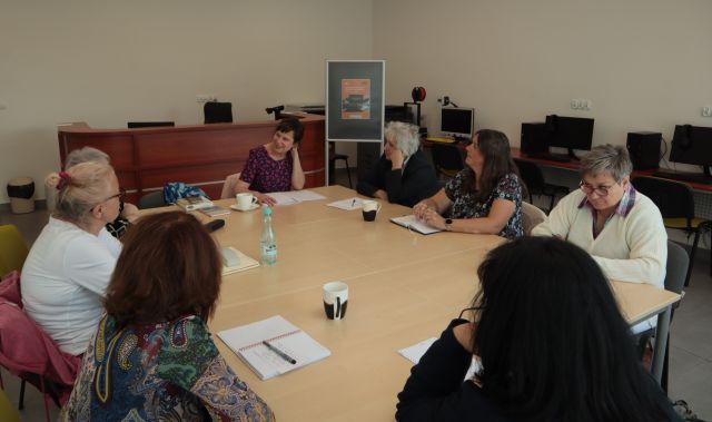 8 seniorek i prowadząca warsztaty siedzą przy stołach ustawionych w prostokąt w sali komputerowej, na stołach leżą kartki i zeszyty , długopisy