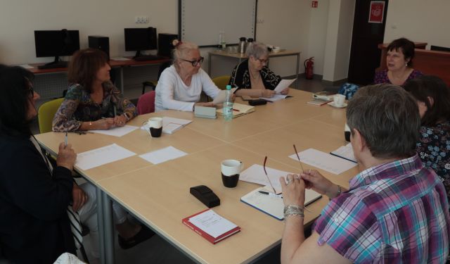 8 seniorek i prowadząca warsztaty siedzą przy stołach ustawionych w prostokąt w sali komputerowej, na stołach leżą kartki i zeszyty , długopisy