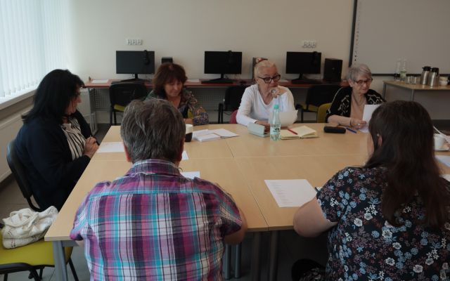 8 seniorek i prowadząca warsztaty siedzą przy stołach ustawionych w prostokąt w sali komputerowej, na stołach leżą kartki i zeszyty , długopisy