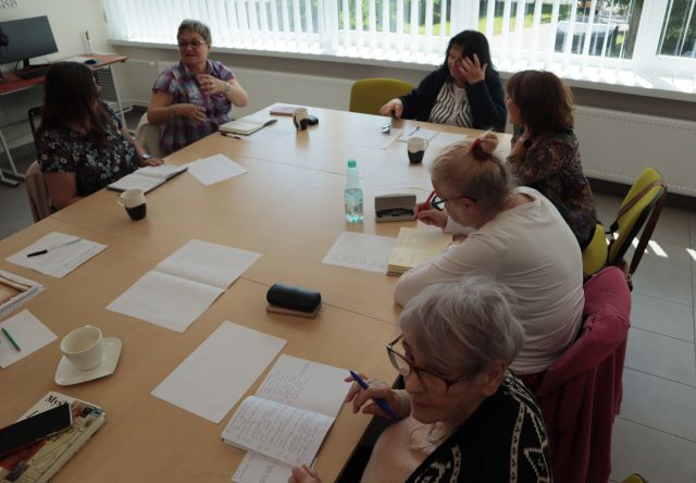 8 seniorek i prowadząca warsztaty siedzą przy stołach ustawionych w prostokąt w sali komputerowej, na stołach leżą kartki i zeszyty , długopisy