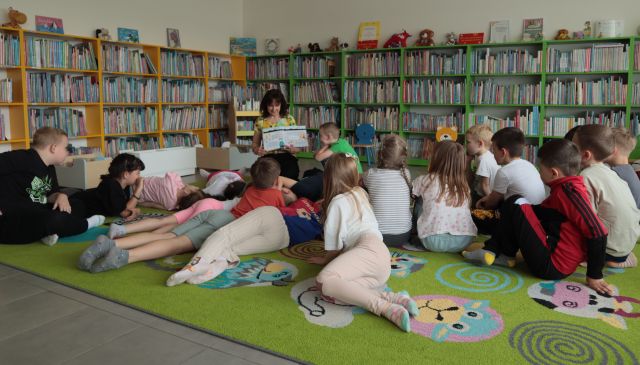 dyrektorka biblioteki siedzi z książką na dywanie, wokół niej na dywanie siedzą i leżą dzieci, w tle regały z książkami