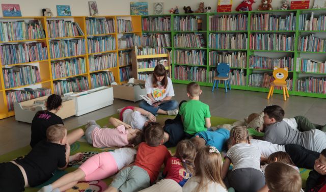 bibliotekarka siedzi z książką na dywanie, wokół niej na dywanie siedzą i leżą dzieci, w tle regały z książkam