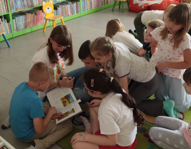bibliotekarka siedzi na dywanie, wokół niej na dywanie siedzą i leżą dzieci, chłopiec czyta książkę, w tle regały z książkami