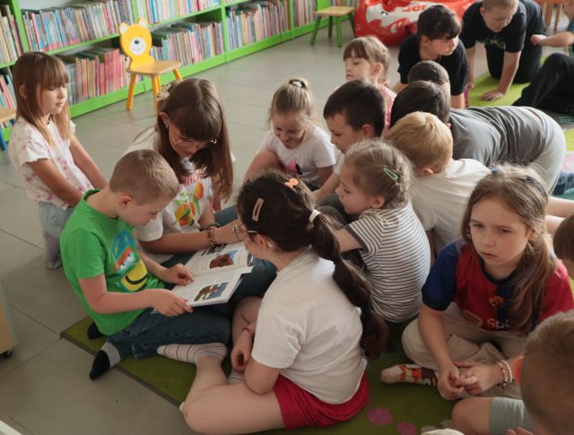 bibliotekarka siedzi na dywanie, wokół niej na dywanie siedzą i leżą dzieci, chłopiec czyta książkę, w tle regały z książkami