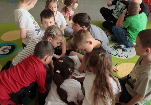 bibliotekarka siedzi na dywanie, wokół niej na dywanie siedzą i leżą dzieci, chłopiec czyta książkę, w tle regały z książkami