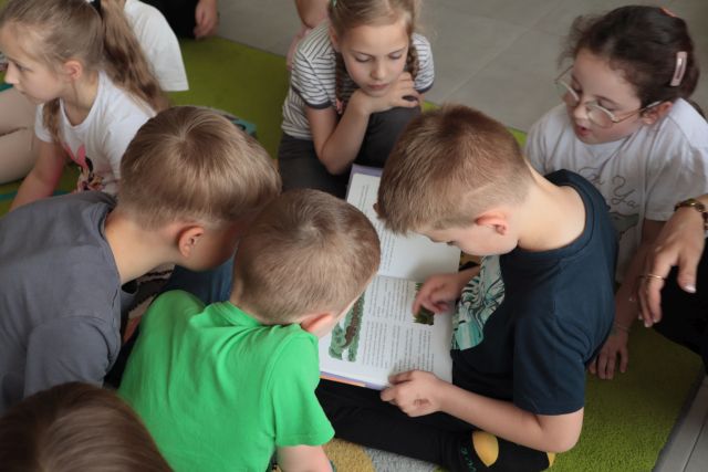 bibliotekarka siedzi na dywanie, wokół niej na dywanie siedzą i leżą dzieci, chłopiec czyta książkę, w tle regały z książkami