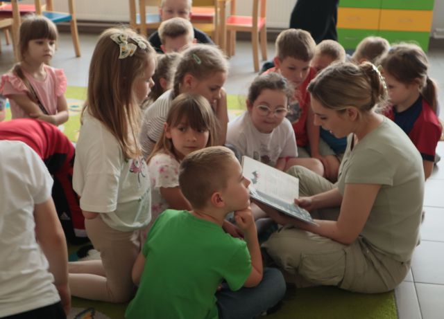 bibliotekarka siedzi na dywanie i czyta, wokół niej na dywanie siedzą i leżą dzieci, w tle regały z książkami