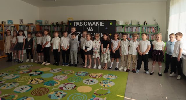 przedszkolaki stoją w rzędzie, w tle tablica z napisem Pasowanie na Czytelnika oraz regały z książkami