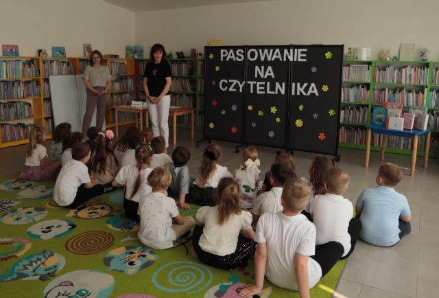 dyrektorka biblioteki stoi przed siedzącymi przedszkolakami, w tle regały z książkami
