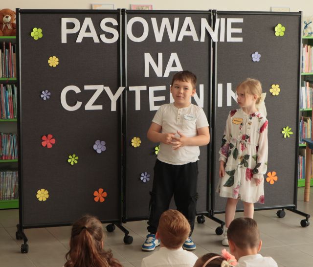 chłopiec i dziewczynka stoją przed tablicą z napisem Pasowanie na Czytelnika
