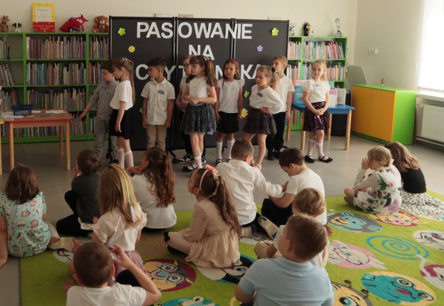 część przedszkolaków siedzi na kolorowym dywanie, a część stoi w kolejce do konkursu