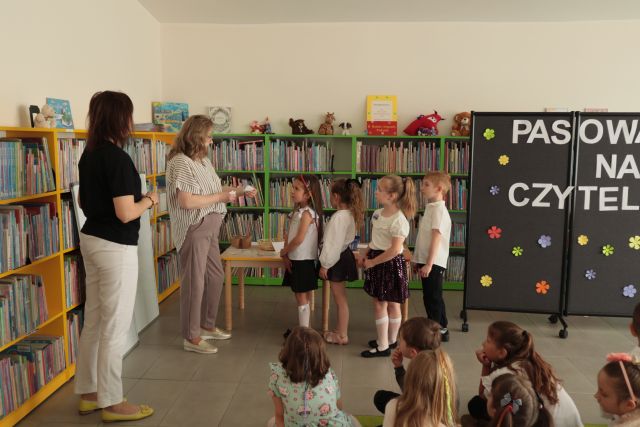 przedszkolaki stoją przed bibliotekarką z kartkami, w tlle regały z książkami
