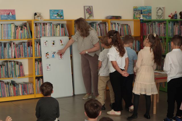 dzieci stoją przed bibliotekarką przypinającą kolorowe ilustracje na tablicy magnetycznej