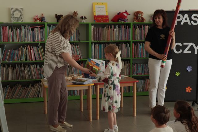 dziewczynka odbiera książkę i dyplom od dyrektorki biblioteki i bibliotekarki