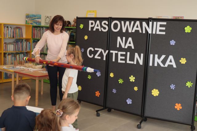 dyrektorka biblioteki pasuje na czytelnika biblioteki ogromnym ołówkiem dziewczynkę