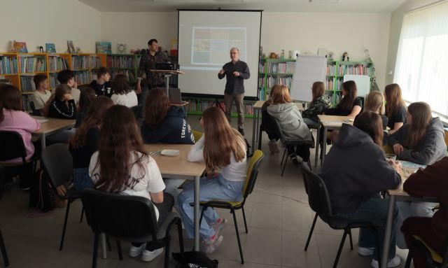 młodzież siedzi przy stolikach, przed nimi stoi dwóch mężczyzn, w tle regały z książkami